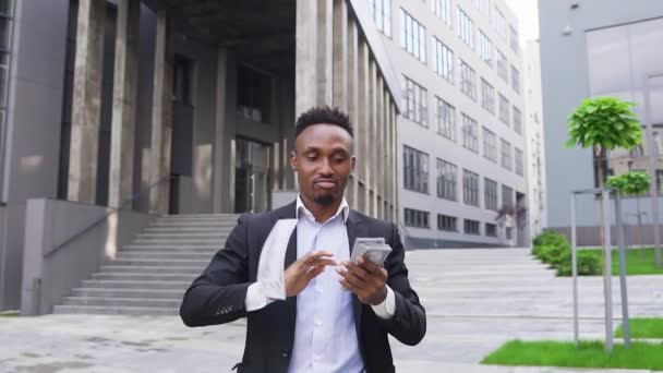 Vista frontal de feliz rico guapo barbudo afroamericano que dispersa dinero en frente de edificio moderno de la ciudad bailando simultáneamente — Vídeo de stock