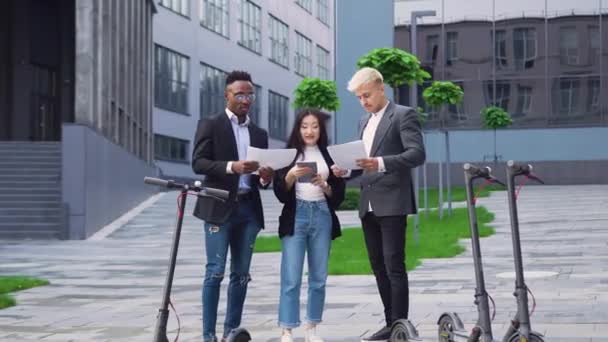 Good-looking confident creative diverse team standing near their e-scooters in the middle of the office buildings yard and browsing financial documents — Stock Video