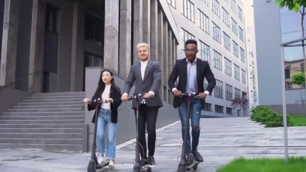 Beau jeune satisfait mixte race amis élégants chevauchant sur leurs propres e-scooters près de l'immeuble de bureaux moderne, 4k — Video