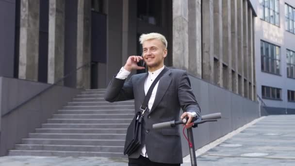 スタイリッシュな服を着た笑顔の幸せな自信に満ちた軽い髪の男市庁舎の近くの電子スクーターで歩くとモバイルで話す — ストック動画