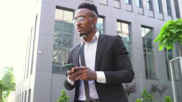 Przystojny, skoncentrowany młody, afrykański Amerykanin w stylowym ubraniu stojący w pobliżu biurowca i korzystający z telefonu, 4k — Wideo stockowe