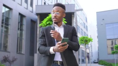 İyi huylu, gülümseyen, 30 yaşında, siyah tenli ofis yöneticisi modern ofis binasının yakınındaki bir sokakta akıllı telefondan video görüşmesi yapıyor.