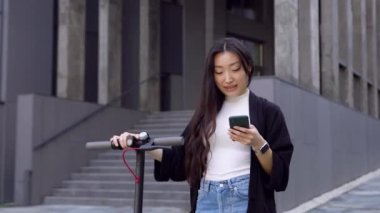 Güzel gülümseyen siyah saçlı Asyalı kız modern binanın yakınında kişisel e-scooterıyla yürüyor ve akıllı telefonunu kullanıyor, 4K