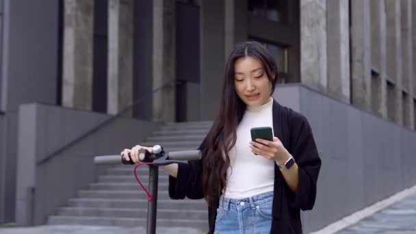 漂亮的黑发亚洲女孩，带着个人电子摩托车走在现代建筑附近，用她的智能手机4k — 图库视频影像