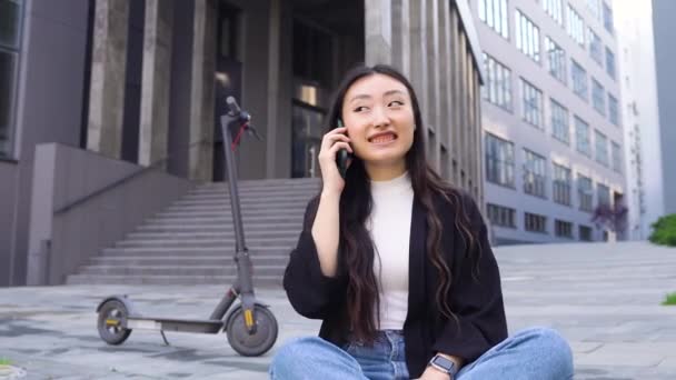 Close up of attractive happy smiling young modern asian girl which having mobile conversation, sitting on concrete ground near urban building, 4k — стоковое видео