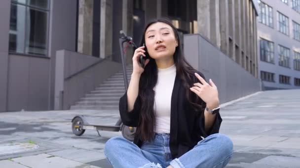 Aangename vrolijke stijlvolle jonge aziatische meisje zitten op asfalt in de buurt van moderne kantoorgebouw en praten op smartphone, 4k — Stockvideo