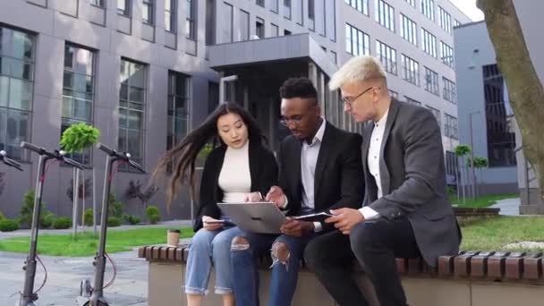 Gut aussehende selbstbewusste, freundliche junge multiethnische Büromenschen, die auf der Bank in der Nähe des Stadtgebäudes sitzen und mit Computern und Finanzberichten zusammenarbeiten — Stockvideo