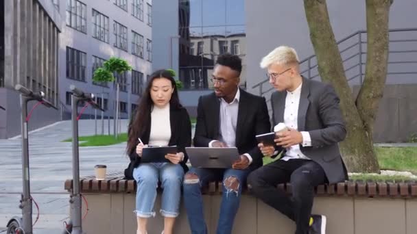Vue de face de sympathique femme asiatique ciblée, les hommes afro-américains et euroréens assis sur le banc près de leur immeuble de bureaux et l'analyse des détails du projet commun — Video