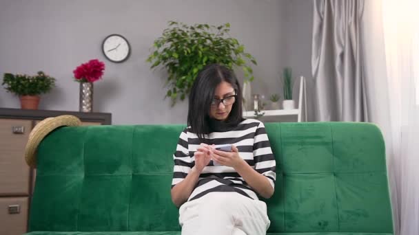 Очаровательная современная молодая женщина в очках и в стильной одежде сидит дома на удобном диване и пользуется смартфоном — стоковое видео