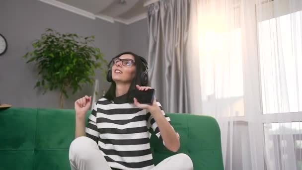 Привлекательная улыбающаяся счастливая молодая темноволосая женщина в наушниках сидит дома на мягком диване и наслаждается современной музыкой из плейлиста телефона — стоковое видео