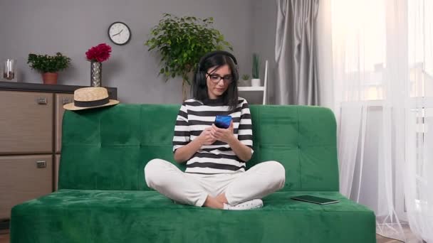 Довольная счастливая современная молодая женщина в наушниках сидит на мягком диване в специально отведенной комнате и слушает приятные песни — стоковое видео
