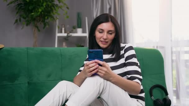 Mujer joven satisfecha sonriente atractiva con cabello oscuro sentada en un sofá verde cerca de la ventana y escribiendo un mensaje a alguien en su teléfono — Vídeos de Stock