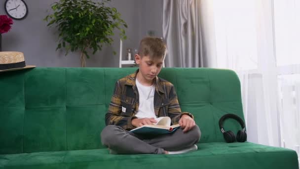 Guapo concentrado adolescente chico está cansado de leer un libro sin interés, sentado en verde sofá suave en casa — Vídeos de Stock