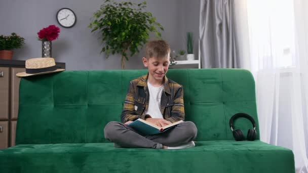 英俊、快乐的现代男孩坐在漂亮设计的房间里舒适的沙发上，听着有趣的书的故事笑了起来 — 图库视频影像