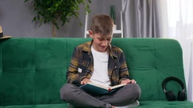 Yakışıklı, neşeli, neşeli, 10 yaşında, evinde yumuşak koltukta komik kitaplar okuyan bir çocuk.