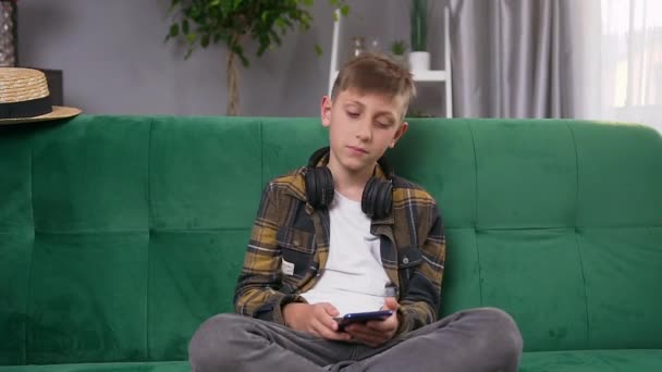 Przystojny, uśmiechnięty, nowoczesny nastolatek ze słuchawkami na szyi, szukający ulubionej piosenki w telefonie, siedząc na kanapie w domu — Wideo stockowe