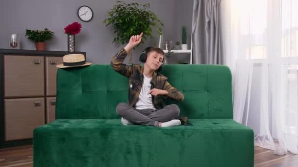 Funny handsome cheerful teen boy sitting on sofa at home and enjoying inflammatory music in headphones with raised hands — Stock Video