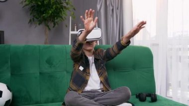 Yumuşak yeşil koltukta oturan ve ilginç bir video oyunu oynamak için fütüristik 3D gözlük kullanan sempatik, çağdaş bir genç çocuk.