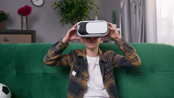 Framifrån av leende stilig smart 10-årig pojke som sitter på soffan och använder virtuell verklighet headset för att spela TV-spel — Stockvideo