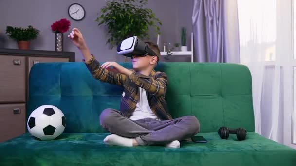 Possível concentrado moderno menino de 10 anos sentado em sofá confortável na sala de estar e brincando na tela imaginária em fone de ouvido de realidade virtual — Vídeo de Stock