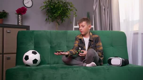 Widok z przodu atrakcyjny pewny siebie inteligentny chłopiec, który siedzi na miękkiej kanapie w domu i trzyma na ręce mały dron i uruchomienie go latać w pokoju — Wideo stockowe