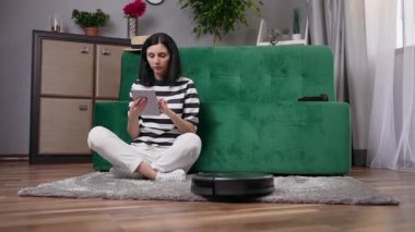 Çekici, kendine güvenen, koltuğun yanında oturan ve robot elektrik süpürgesini kontrol etmek için tablet bilgisayar kullanan genç bayanı kapatın.