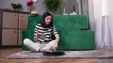 Sevimli odaklı şık esmer kanepenin yanında oturuyor ve i-pad üzerinde talimatlar kullanarak robot süpürge uyguluyor.