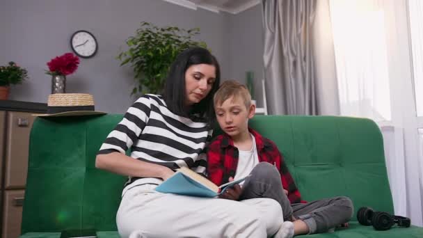 Привабливий задоволений усміхнений безтурботний мати і син читають книгу разом, сидячи на зручному дивані в сучасній кімнаті, повільний рух — стокове відео