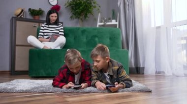 Mutlu gülümseyen güzel annenin kanepeye uzandığı ve iki yakışıklı oğlunun yerde yatıp akıllı telefonlar kullandığı bir aile.