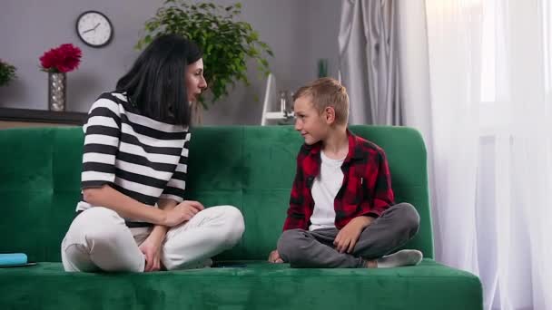Привлекательная сердитая молодая мать ругает своего непослушного 8-летнего сына, и он демонстративно отворачивается, сидя вместе на диване дома — стоковое видео
