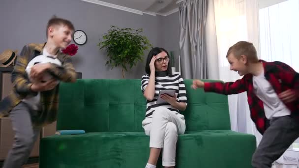 Ελκυστική συγκεντρωμένη πολυάσχολη νεαρή γυναίκα σε γυαλιά που εργάζονται με tablet PC και αίσθημα πονοκέφαλου becouse θορυβώδη έφηβος γιους της τρέχει γύρω από τον καναπέ για να κάθεται — Αρχείο Βίντεο