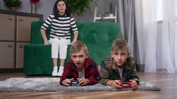 Beautiful happy young woman passing by her handsome positive smiling sons which lying on the floor and playing video game using joysticks — Stock Video