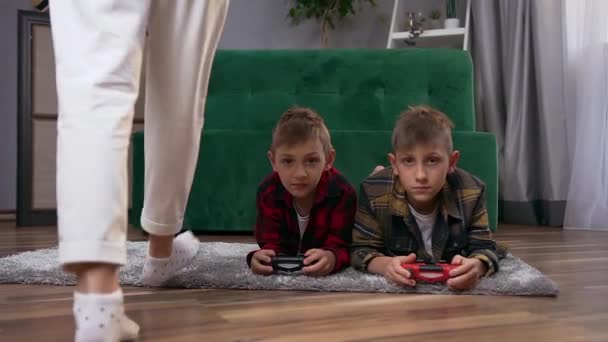 Provável sorte positivo adolescente irmãos deitado no chão e jogar jogos de vídeo usando gamepads quando o seu atraente feliz jovem mãe caming no quarto e sentar-se no sofá — Vídeo de Stock