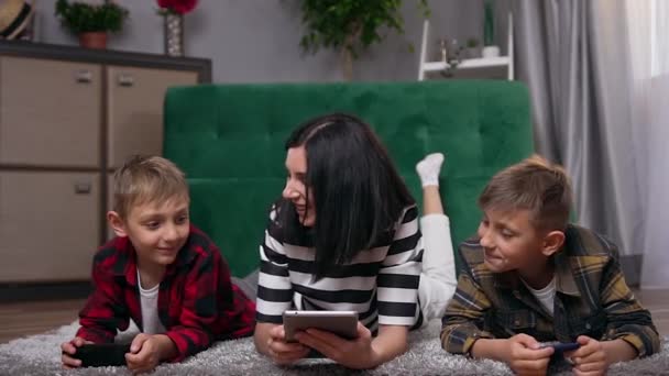 Ritratto di bella sorridente madre amichevole felice ei suoi due figli che sdraiati insieme sul pavimento e utilizzando gadget moderni — Video Stock