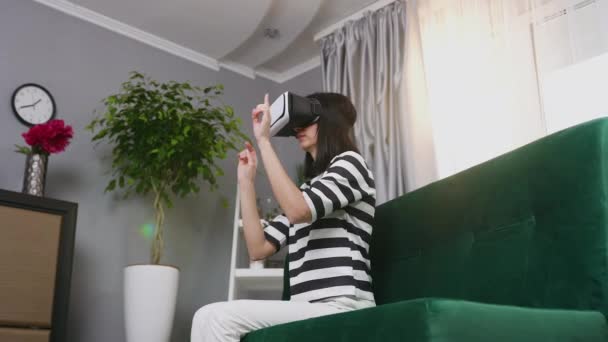 Vue latérale d'une jolie jeune femme moderne concentrée dans des lunettes de réalité augmentée qui s'assoient sur un canapé confortable à la maison et travaillent sur un écran imaginaire, 4k — Video