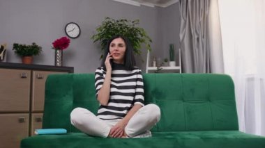 Güzel, gülümseyen, siyah saçlı, şık elbiseli, rahat bir şekilde koltukta oturan ve telefonda konuşan genç bir kadın.