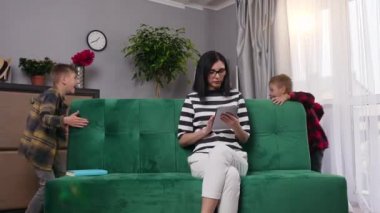 Gözlüklü, konsantre olmuş genç bir kadının tabletle çalışması, gürültücü iki oğlun ona müdahale etmesi ve üzerine oturduğu kanepenin etrafında koşması, 4k
