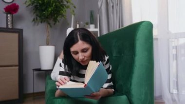 Evdeki yumuşak kanepede uzanıp kitap okuyan, gamsız, gamsız, siyah saçlı genç bir kadını yakından çek.