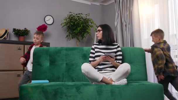 Krásná unavená mladá matka sedí na pohovce v obývacím pokoji a hádají se její dva hravé hlučné syny, které pobíhají po pohovce, 4k — Stock video