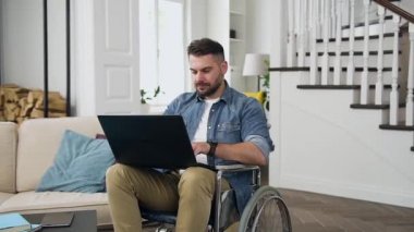 Çekici dengeli sakallı, şık sakallı genç adam tekerlekli sandalyede oturuyor ve evde dizüstü bilgisayarla çalışıyor.