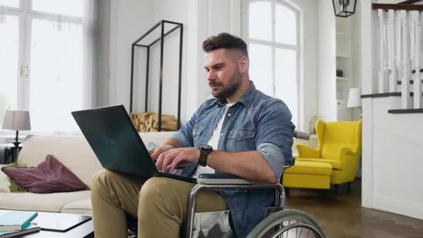 Hombre moderno altamente cualificado y concentrado agradable con barba sentada en silla de ruedas y trabajando remotamente en casa en el portátil — Vídeos de Stock