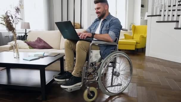 Guapo sonriente satisfecho joven barbudo en silla de ruedas trabajando remotamente en el ordenador personal en casa — Vídeo de stock