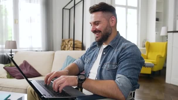 Gros plan de beau jeune homme barbu joyeux et moderne en vêtements décontractés assis en fauteuil roulant à la maison et travaillant à distance sur un ordinateur portable — Video