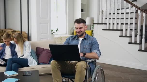 Семейная концепция красивого счастливого улыбающегося бородатого парня в инвалидной коляске, работающего на ноутбуке, а его жена и сын читают книгу на диване рядом с ним — стоковое видео
