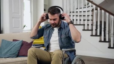 Tekerlekli sandalyedeki mutlu sakallı, çekici genç adamın ön görüntüsü. Kulaklık takıyor ve evde rahatlatıcı müzik dinliyor.