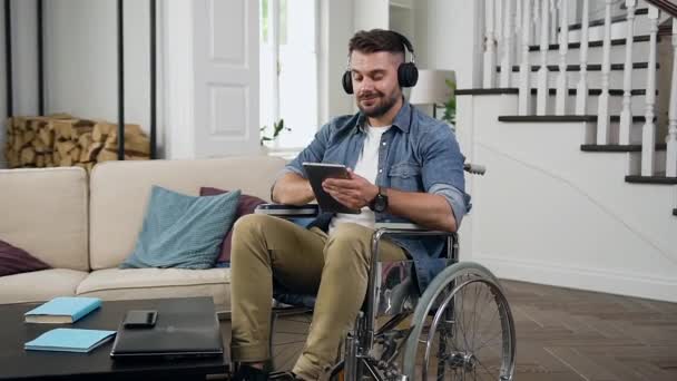 Bello rilassato sorridente ragazzo barbuto in cuffia seduto in sedia a rotelle in soggiorno e ascoltare musica da i-pad — Video Stock