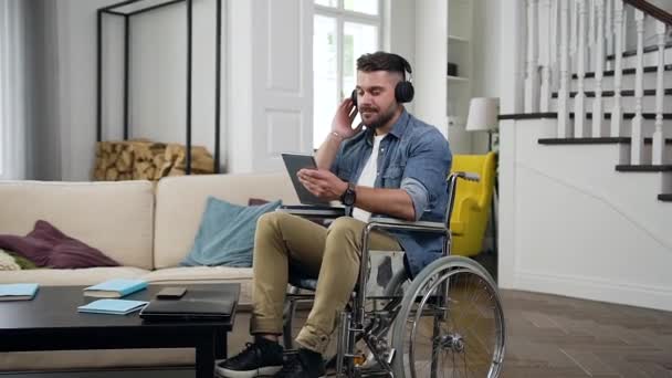 Закрыть положительного счастливчика молодого бородатого парня в инвалидной коляске, который весело проводит время в одиночестве, наслаждаясь танцевальной музыкой в наушниках дома — стоковое видео