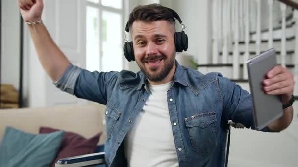 Jeune homme émotionnel joyeux et de bonne humeur avec la barbe assise en fauteuil roulant et dansant sous la musique inflammatoire dans les écouteurs à la maison — Video