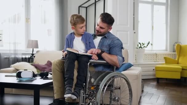 Familie concept van goedgehumeurde gelukkige jonge bebaarde gehandicapte vader in een rolstoel die helpt om boek te lezen aan zijn kleine zoon die zit op zijn knieën — Stockvideo