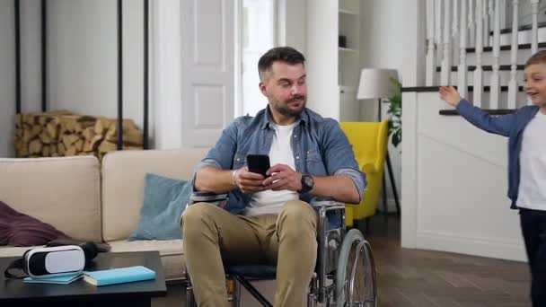Доброзичливий усміхнений 30-річний бородатий чоловік у інвалідному візку використовує свій смартфон і обіймає свого веселого маленького сина, який біжить до нього — стокове відео